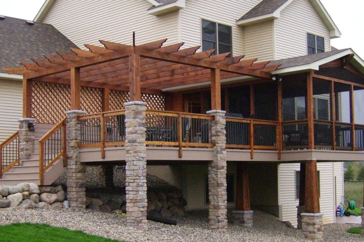 A large, elevated deck featuring a substantial wooden pergola with stone columns, attached to a beige-sided house with a gravel and rock landscape below.