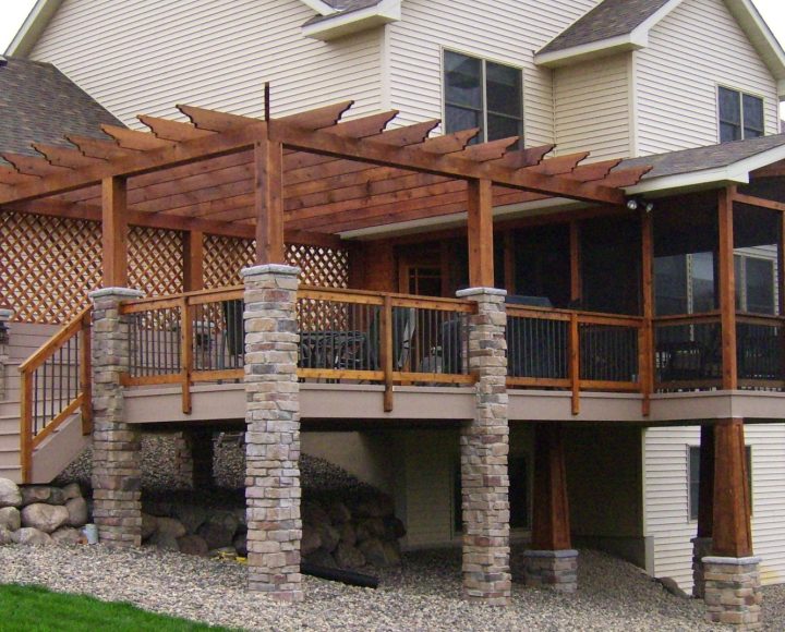 A large, elevated deck featuring a substantial wooden pergola with stone columns, attached to a beige-sided house with a gravel and rock landscape below.