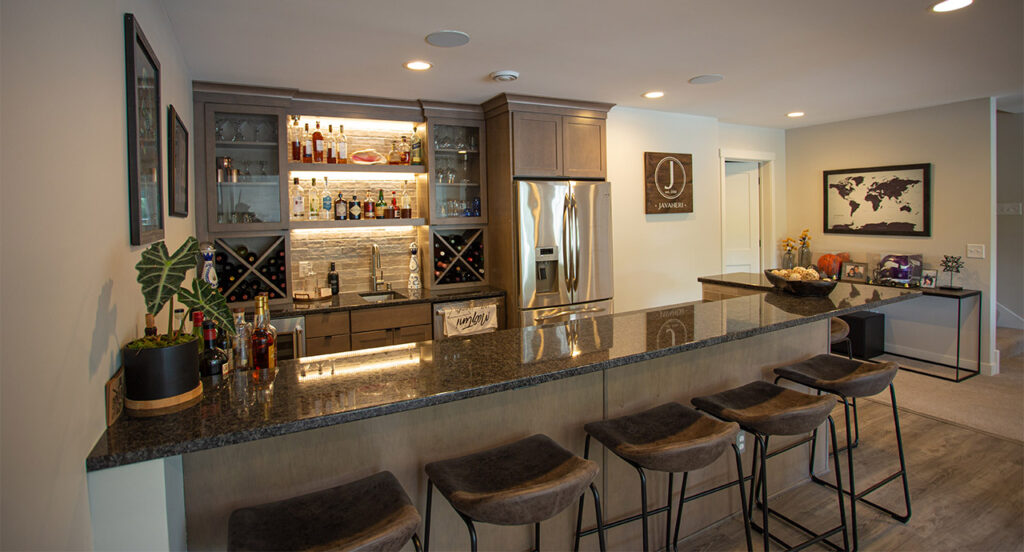 A sleek home bar with a granite countertop, backlit shelves displaying bottles, wine storage, a stainless steel refrigerator, and bar stools.