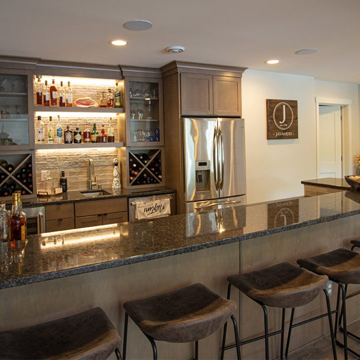 A sleek home bar with a granite countertop, backlit shelves displaying bottles, wine storage, a stainless steel refrigerator, and bar stools.