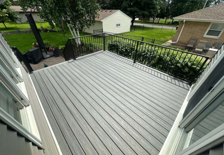 A large, empty gray composite deck with black metal railings, viewed from above, overlooking a green backyard with trees and neighboring houses.