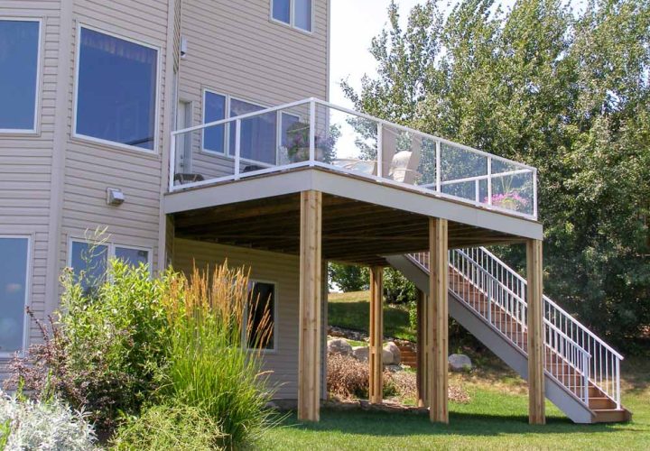 A two-tiered deck with white posts and glass railings attached to a beige-sided house, situated on a landscaped yard with ornamental grasses and trees.