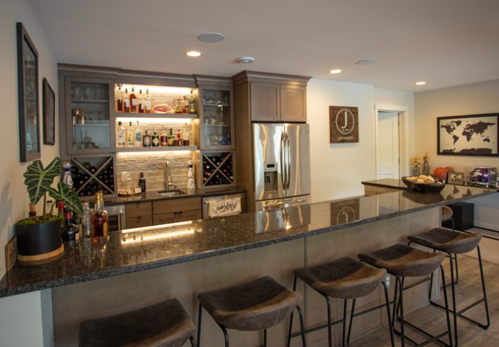 A modern home bar with backlit shelves displaying bottles, a stainless steel refrigerator, a granite countertop, and bar stools.