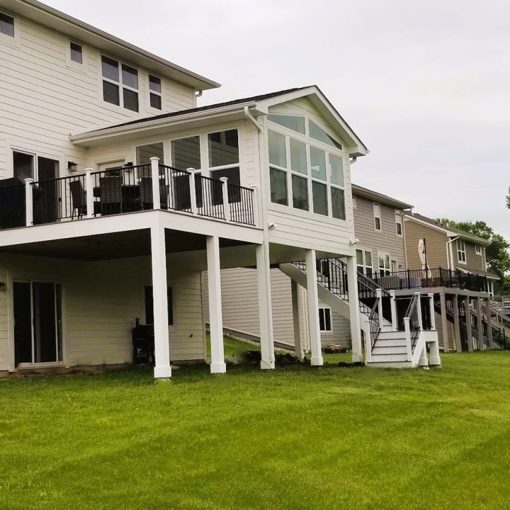 A line of contemporary homes with white siding and large upper decks, each supported by white posts and connected by stairs, overlooking a well-maintained green lawn.