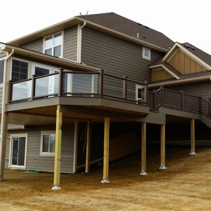 A substantial two-tiered deck with brown railings and stairs attached to a gray-sided house, situated on a sloped, dry grassy yard.