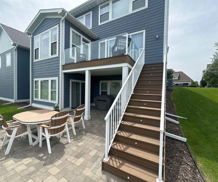 The back of a blue-sided home with a modern glass-railed upper deck and a lower paver patio furnished with a wooden dining table and chairs.
