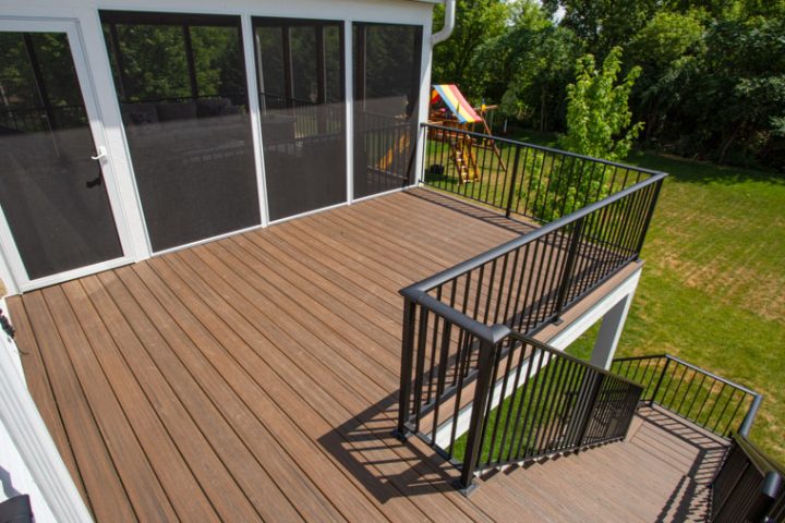 A screened porch with brown composite decking and black railings, featuring a view of a green lawn and a children's playset in the background.