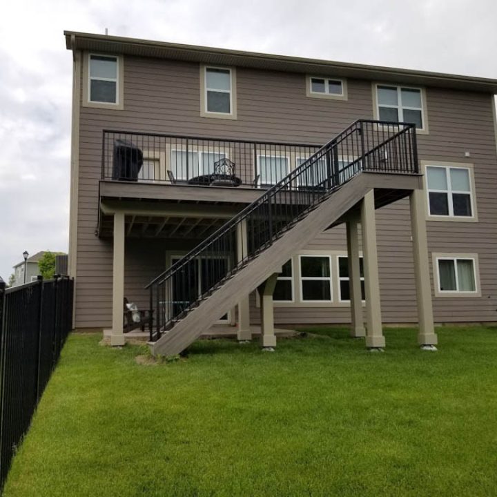A two-tiered deck with black railings and stairs attached to a gray-sided house, set against a backdrop of a green lawn and a black fence.