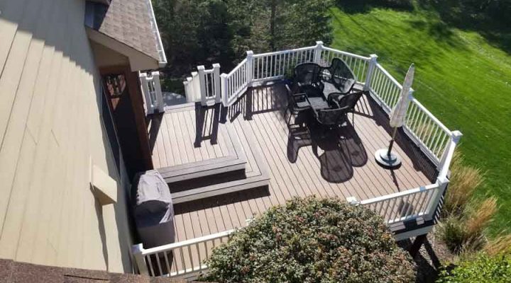 An elevated deck with white railings and stairs, featuring outdoor furniture and an umbrella, overlooking a steep, grassy slope and trees.