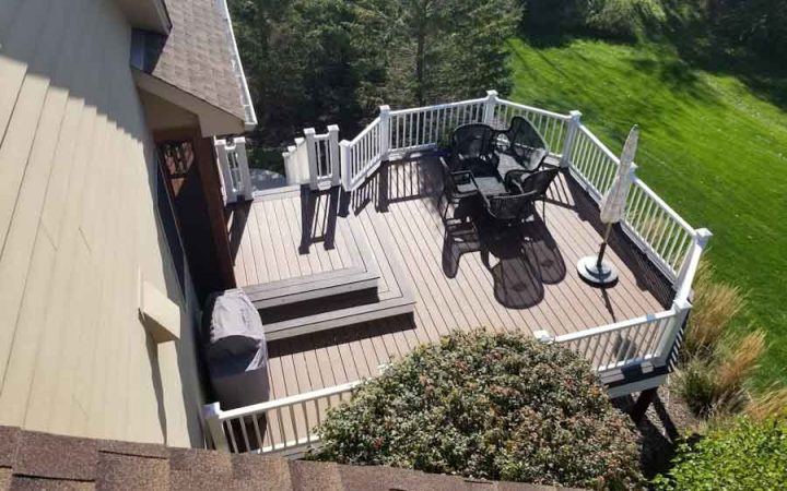 An elevated deck with white railings and stairs, featuring outdoor furniture and an umbrella, overlooking a steep, grassy slope and trees.