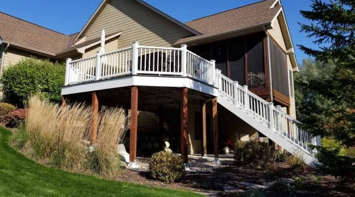 A two-tiered deck with white railings attached to a beige-sided house, situated on a sloped, landscaped yard with ornamental grasses and evergreen shrubs.