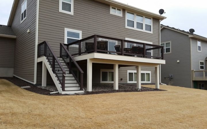 A two-tiered deck with brown railings and stairs attached to a gray-sided house, situated on a sloped, dry grassy yard.