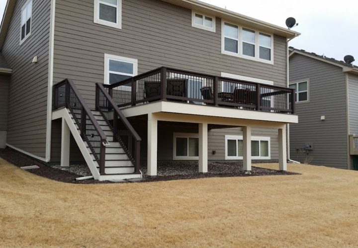 A two-tiered deck with brown railings and stairs attached to a gray-sided house, situated on a sloped, dry grassy yard.