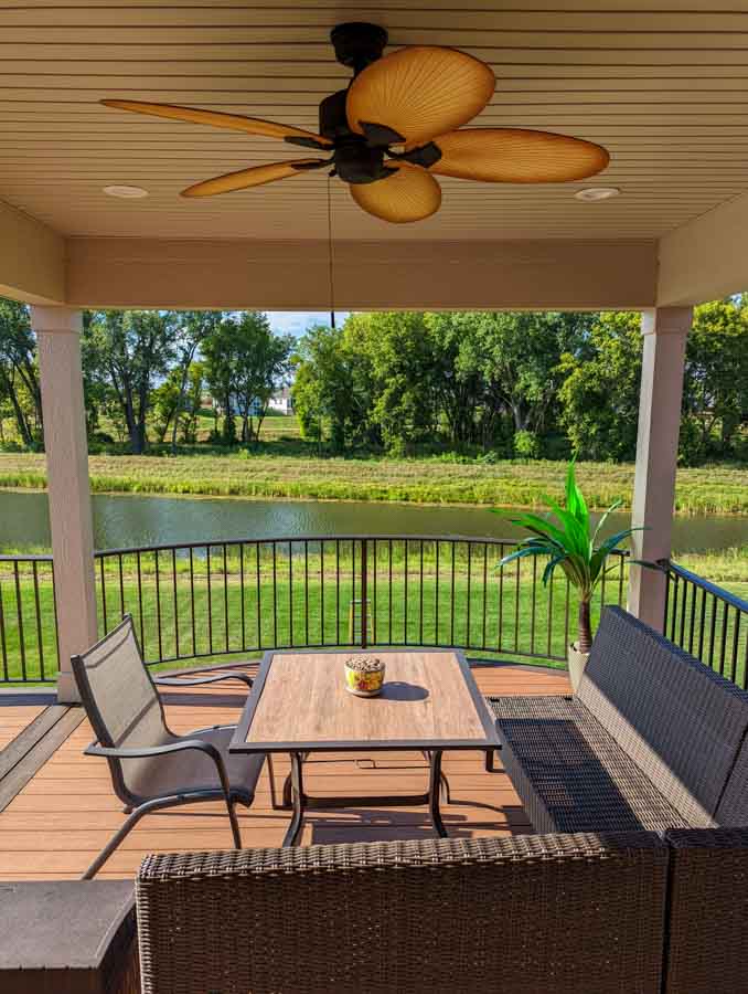 A comfortable wicker patio set on a wooden deck with a view of a tranquil pond and green fields beyond the railing.
