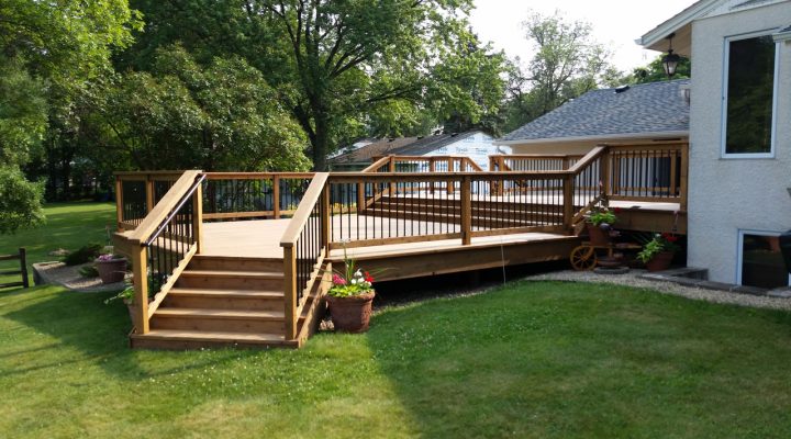 A large, multi-level wooden deck with black metal railings and stairs, surrounded by a lush green lawn and mature trees on a sunny day.