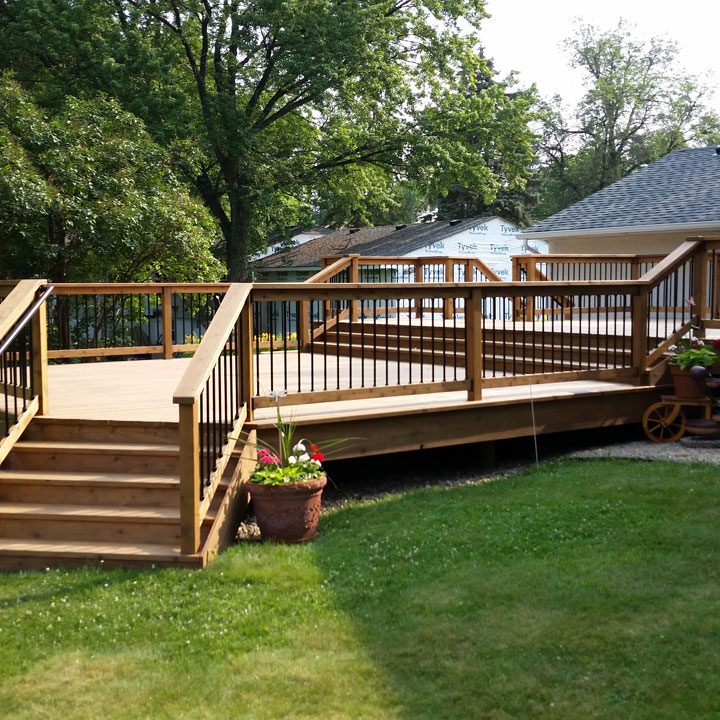A large, multi-level wooden deck with black metal railings and stairs, surrounded by a lush green lawn and mature trees on a sunny day.