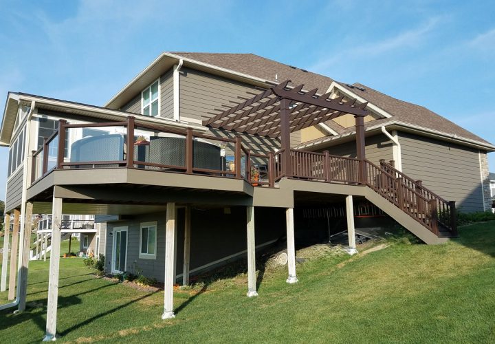 A large, modern deck with brown railings and a pergola attached to a gray-sided house on a sunny day.