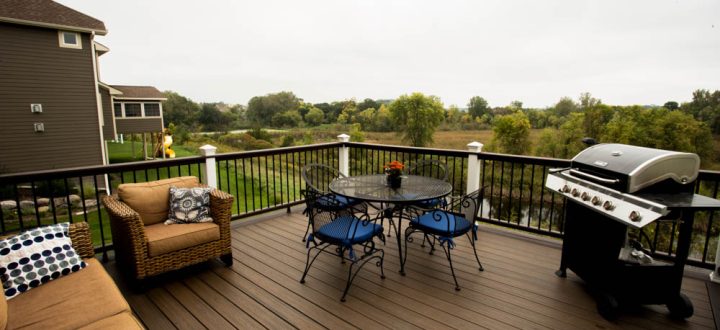 A dark wood deck with wicker furniture, a glass-top table, and a grill, featuring white posts and black railings that overlook a scenic river and green fields.